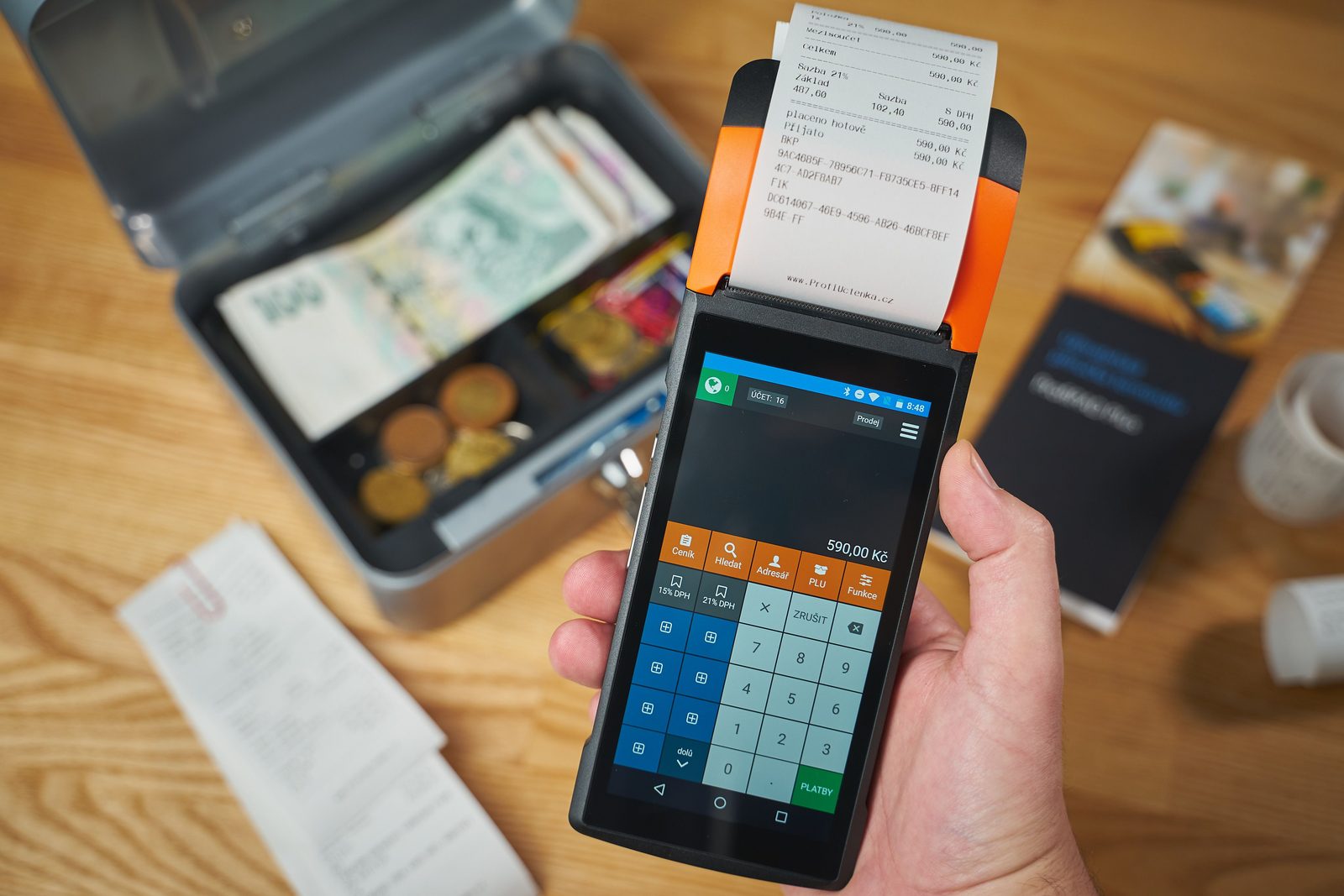 A handheld payment terminal printing a receipt beside an open cash box of banknotes and coins